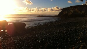 Ein felsiger Strand bei Sonnenuntergang, ruhige See und eine Klippe mit Palmen.