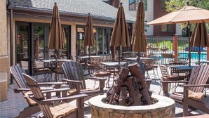 Une terrasse extérieure avec des chaises et des tables en bois, entourée de parasols, et un foyer en pierre au centre.