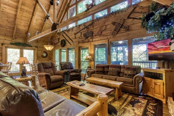 A cozy cabin interior with wooden walls, large windows, a fireplace, leather furniture, and rustic decor.