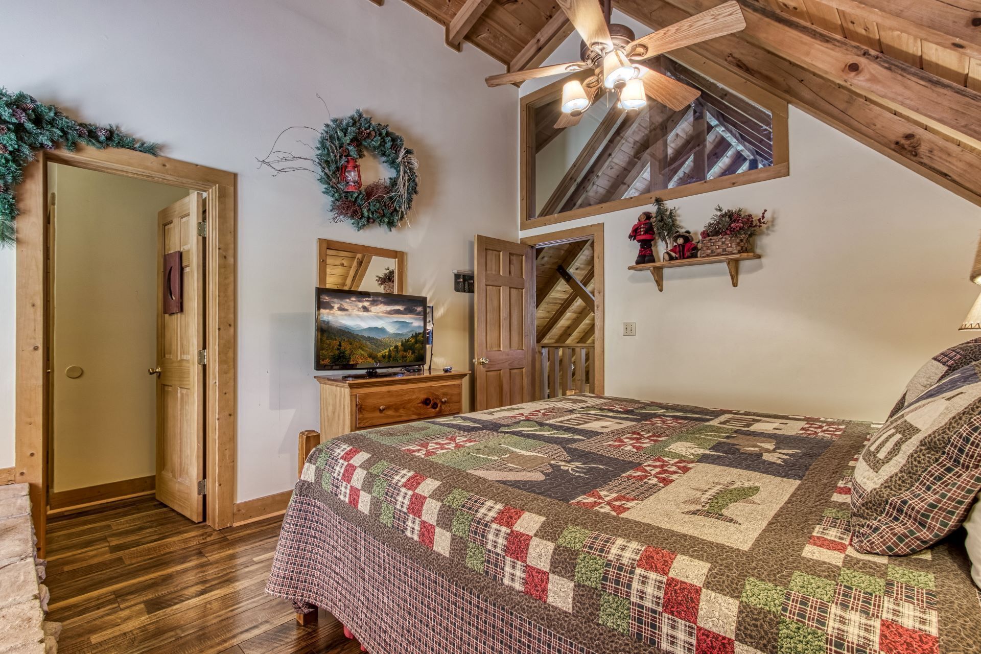 A cozy bedroom with a wooden ceiling, a large bed with a quilt, a television, and a mirror.