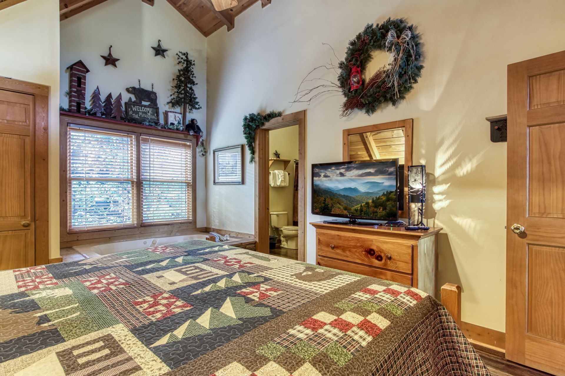 A cozy bedroom with a large bed, a television, a wooden dresser, and a window with blinds.