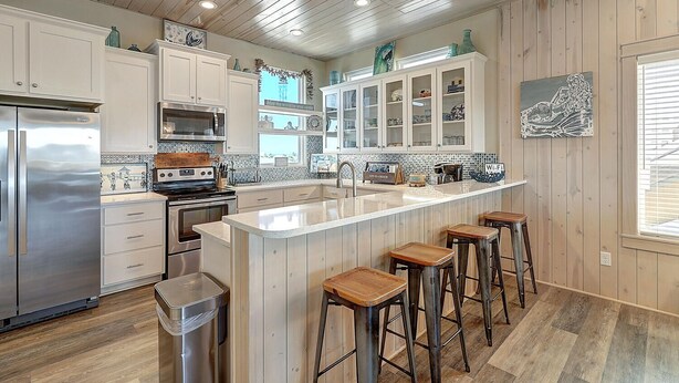 A modern kitchen with white cabinets, a stainless steel refrigerator, a central island with bar stools, and wooden flooring. - Detail and craftsmanship are evident in every space. From the designer kitchen a (Port Aransas)