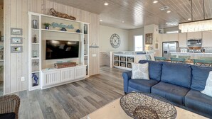 A spacious living room with a large flat-screen TV, a blue sofa, and a dining area with a kitchen in the background. - Detail and craftsmanship are evident in every space. From the designer kitchen a (Port Aransas)