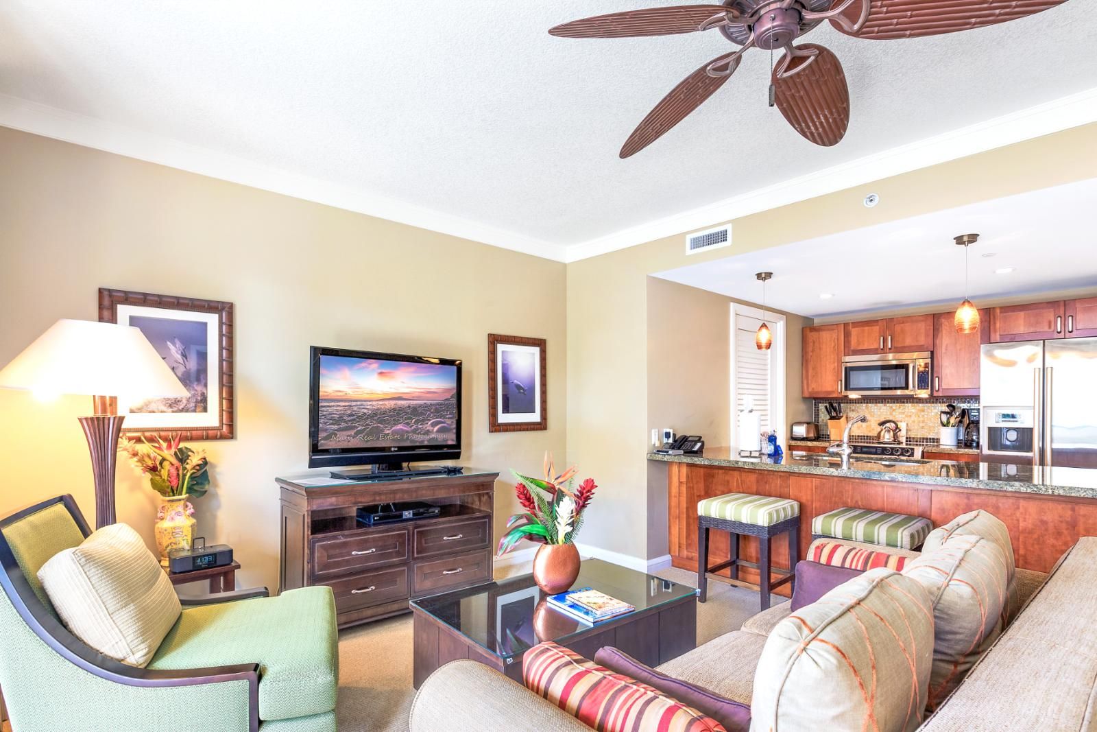 A living room with a TV, sofa, and coffee table. There is a kitchen area with a microwave, refrigerator, and bar stools.