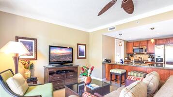 A living room with a TV, sofa, and coffee table. There is a kitchen area with a microwave, refrigerator, and bar stools.
