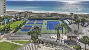 En tennisbane med udsigt til stranden, omgivet af palmer og en bygning.