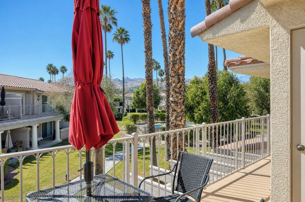 Ein Balkon mit Tisch und Stühlen, einem roten Sonnenschirm und Blick auf einen Pool sowie Palmen.
