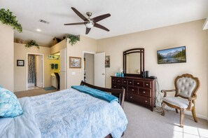 Ein Schlafzimmer mit einem Bett, einem Schminktisch, einem Sessel, einem Deckenventilator und einem Bild an der Wand.