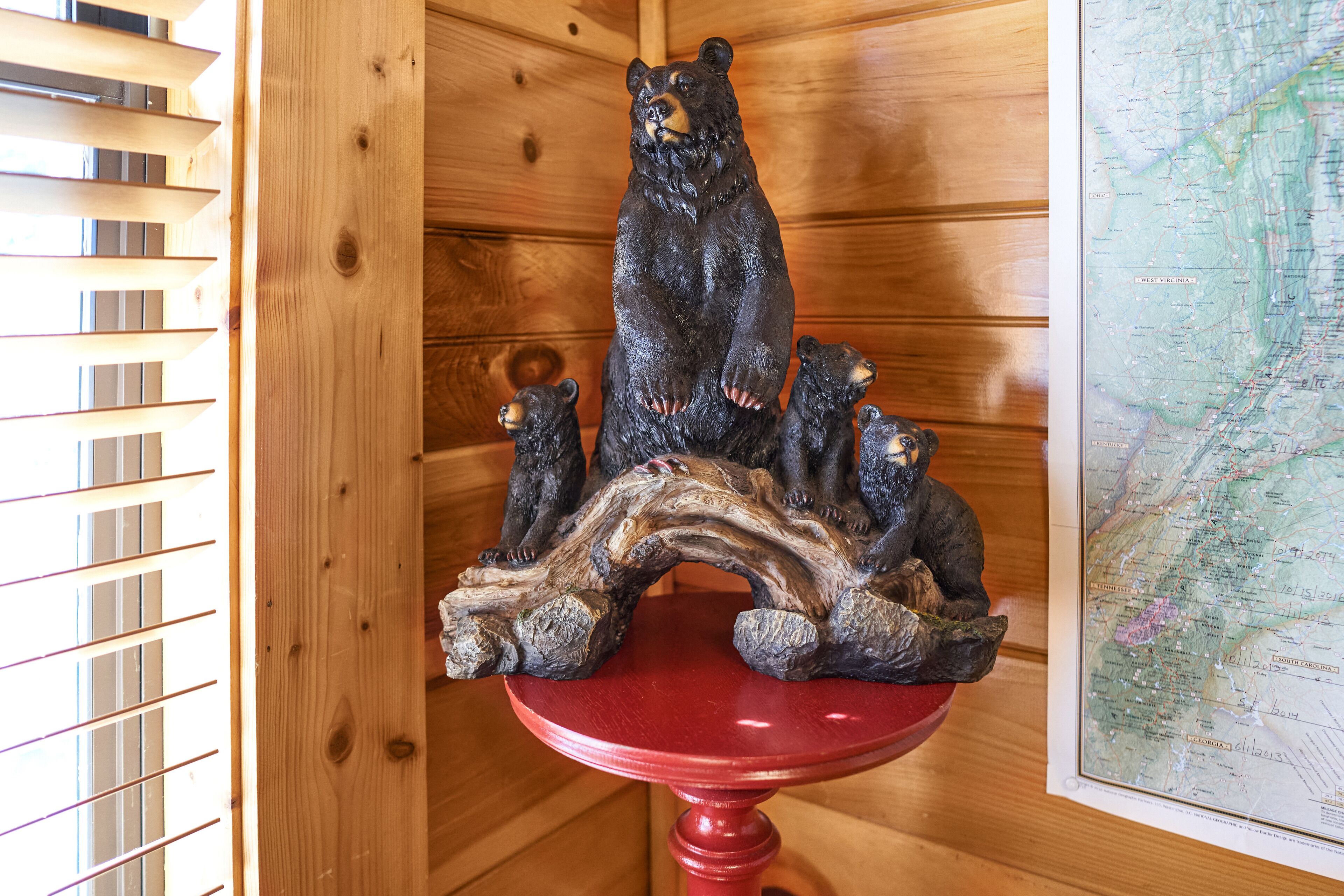 Interior de una cabaña de madera con una escultura de oso de madera sobre un pedestal rojo, un mapa en la pared y listones de madera a la izquierda.