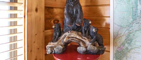 Interior de una cabaña de madera con una escultura de oso de madera sobre un pedestal rojo, un mapa en la pared y listones de madera a la izquierda.