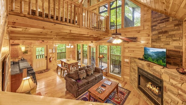 A spacious cabin with a wooden interior, a fireplace, a television, and a balcony overlooking a forested area.