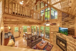 A spacious cabin with a wooden interior, a fireplace, a television, and a balcony overlooking a forested area. - Cherokee Charm: Theater, Gameroom, Hot Tub, Sauna (Gatlinburg)