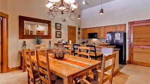 Un comedor con mesa y sillas de madera, una araña y una cocina al fondo.