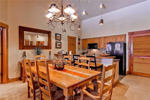 A dining area with a wooden table and chairs, a chandelier, and a kitchen in the background.