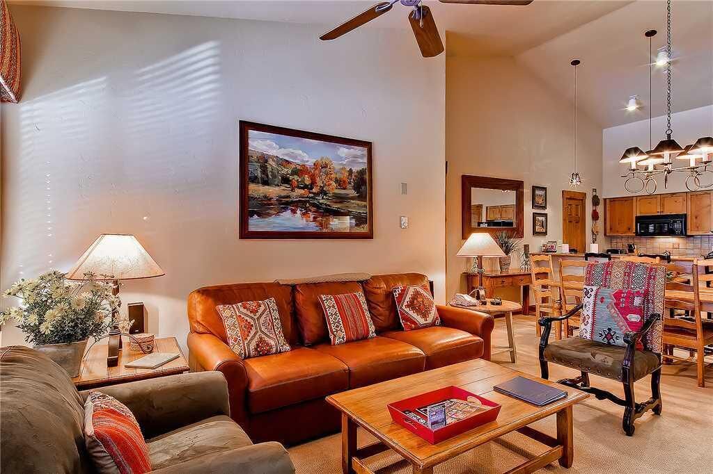 A living room with a brown leather sofa, a wooden coffee table, and a dining area with wooden chairs and a kitchen in the background.