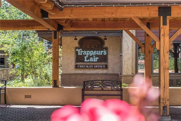 A wooden pergola structure with a sign reading "Trappeur's Lair CHECK-IN OFFICE" in front of a building.