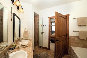 A bathroom with two sinks, a large mirror, and a wooden door leading to a bedroom.