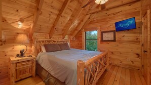 A wooden cabin bedroom with a bed, nightstand, lamp, and a television on the wall.
