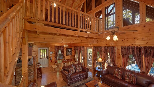 A spacious wooden cabin with a loft bedroom, a cozy living room with leather sofas, and large windows offering views of the outdoors.