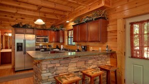 A kitchen with a stone island, wooden cabinets, a refrigerator, an oven, a microwave, and a sink. There are three wooden stools at the island.