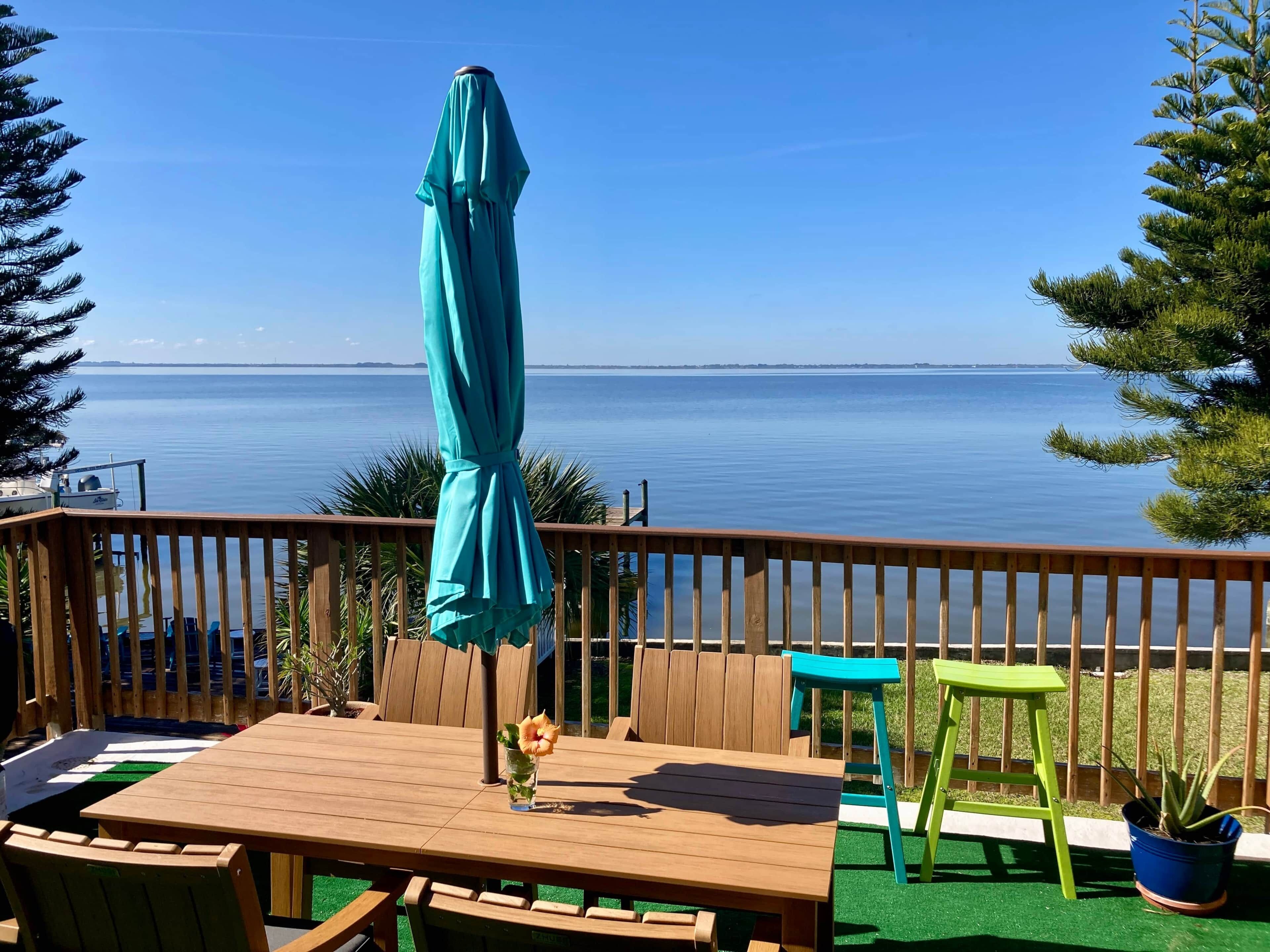 Een houten terras met een tafel, stoelen en een turquoise parasol, met uitzicht op het water.