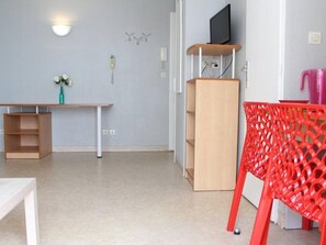 Ein Zimmer mit einem Holztisch, einem roten Sessel, einem Fernseher auf einem Regal und einer Wandvase mit Blumen.