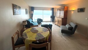 A cozy living room with a sofa, a dining table with chairs, a TV on a stand, and a view of the sea through the window.