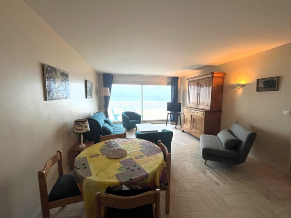 Un salon confortable avec un canapé, une table à manger et des chaises, un téléviseur posé sur un meuble, et une vue sur la mer par la fenêtre.