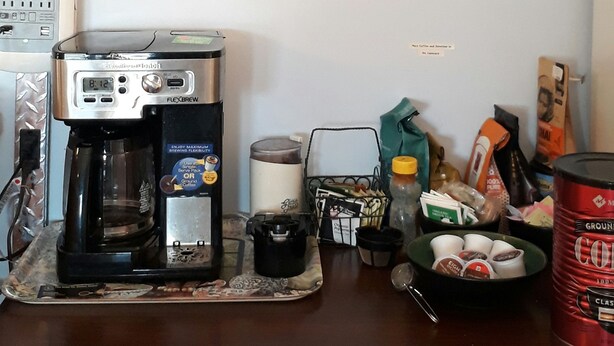 A coffee maker on a counter, a framed picture of a boat on water, and a coffee bag on the wall.