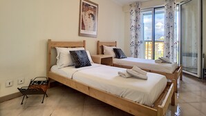 Une chambre d’hôtel avec deux lits en bois, une petite table, une chaise et une grande fenêtre avec des rideaux.