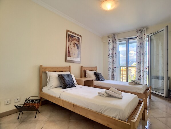 Une chambre d’hôtel avec deux lits en bois, une petite table, une chaise et une grande fenêtre avec des rideaux.