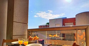 A table set for breakfast with orange juice, pastries, and condiments on a terrace with a view of buildings and a clear sky.