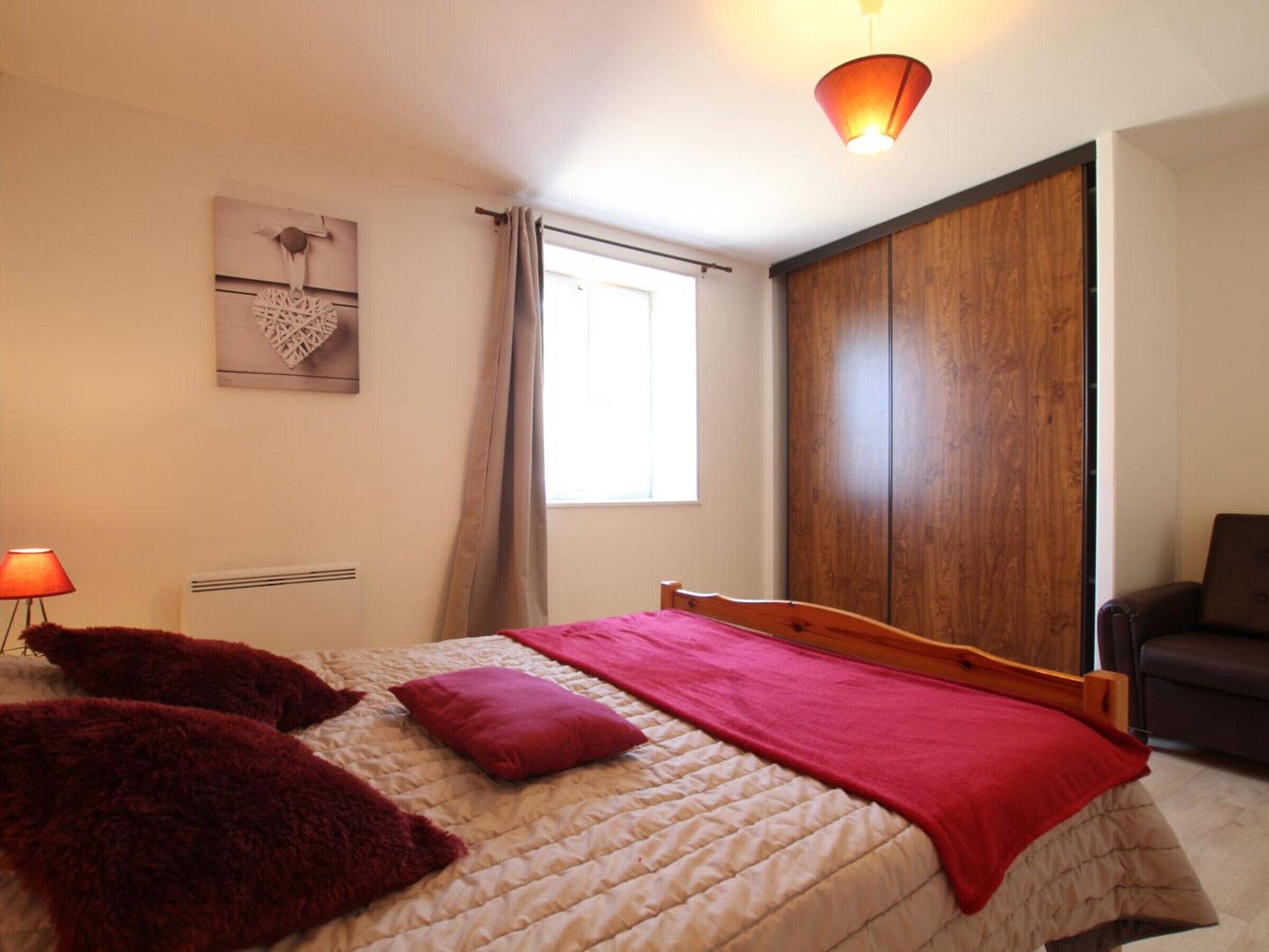 A bedroom with a bed, a wooden wardrobe, a window with curtains, and a lamp.