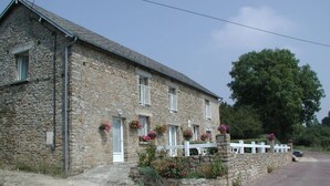 A stone building with a white fence, a gravel driveway, and a large tree in the background.