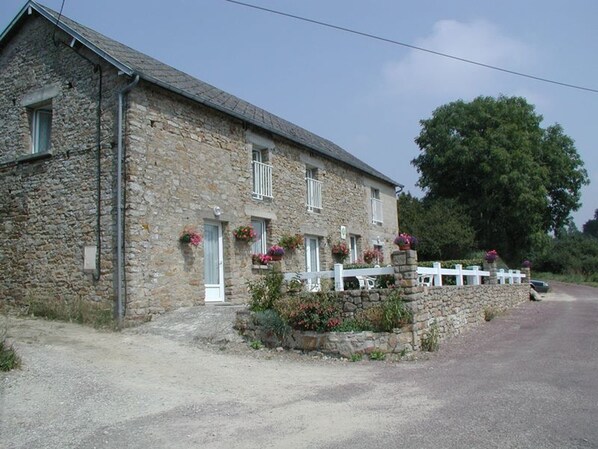 A stone building with a white fence, a gravel driveway, and a large tree in the background.