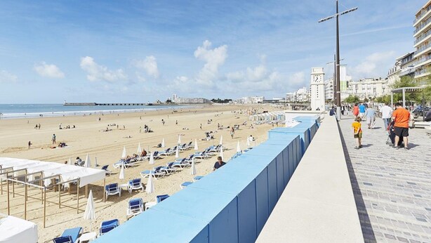 A beach with sun loungers and umbrellas, a promenade with people, and buildings in the background.