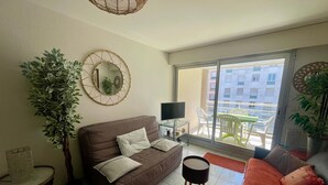 A living room with a brown sofa, a small round table, a TV, and a balcony with outdoor furniture.