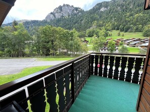 Un balcon avec un tapis vert, une rambarde noire et une vue sur une région montagneuse parsemée de maisons.