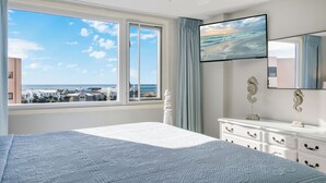 Ein Schlafzimmer mit einem großen Bett, einem Schminktisch, einem Fernseher und Blick auf das Meer und Gebäude.