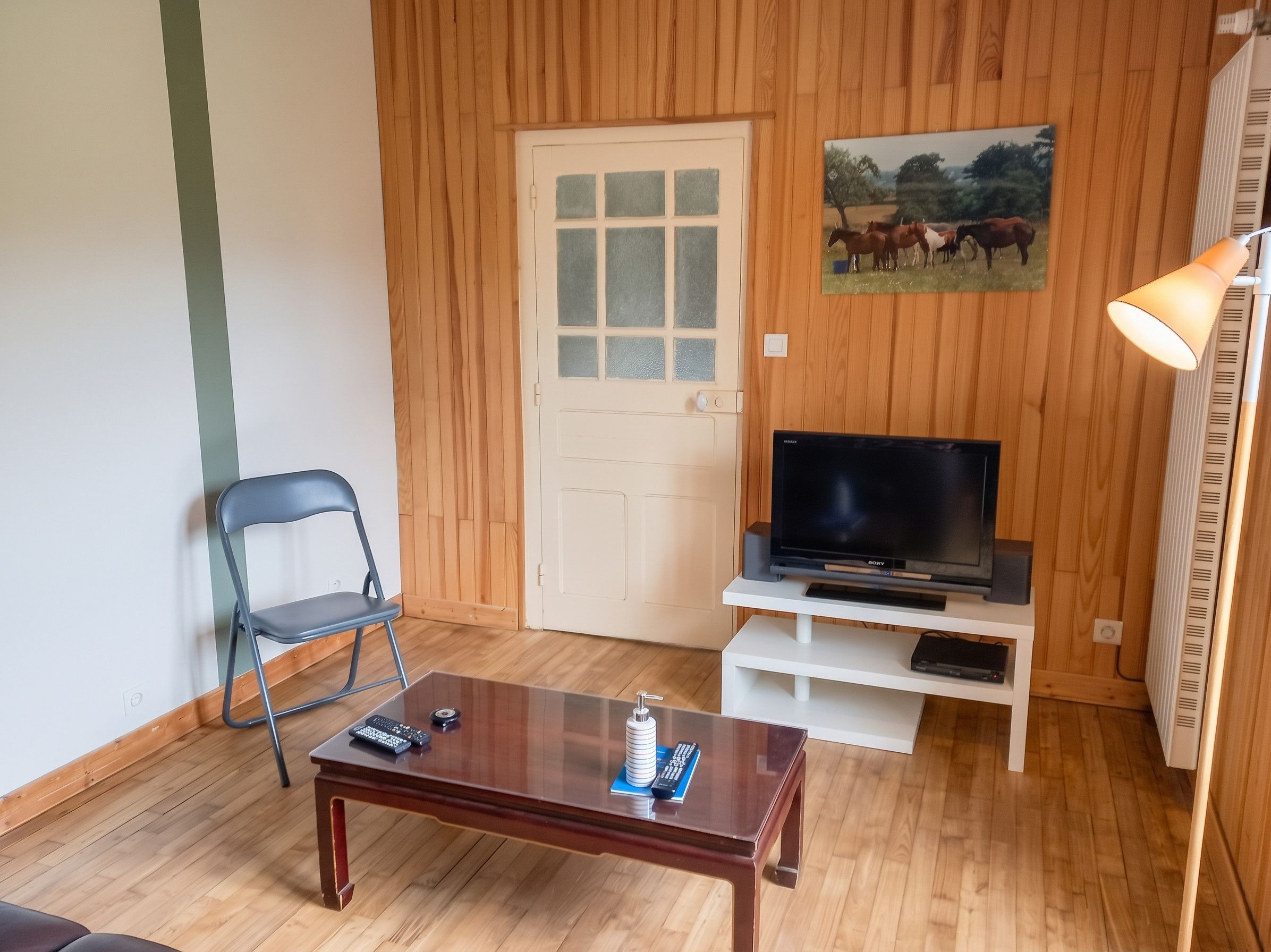 A room with a wooden door, a television on a white stand, a folding chair, and a small table with a remote control.
