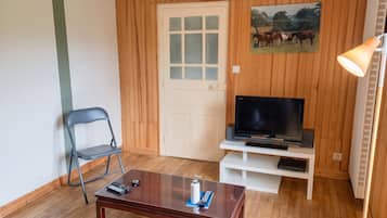 A room with a wooden door, a television on a white stand, a folding chair, and a small table with a remote control.