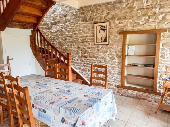 A dining area with a table covered in a nautical-themed tablecloth, wooden chairs, a stone wall, a staircase, and a bookshelf. - Homerez - House in Plehedel (Plehedel)