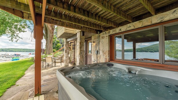Un bain à remous installé sous une pergola en bois, avec des murs en pierre et de grandes fenêtres donnant sur un lac.