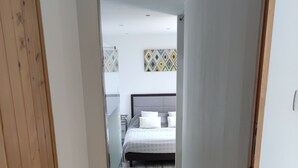 Un couloir étroit mène à une chambre à coucher qui contient un lit, une table de chevet et un luminaire fixé au mur.