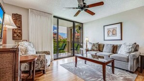 A spacious living room with a wooden floor, a ceiling fan, a sofa set, a coffee table, and a view through a glass door to the outdoors.