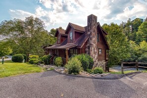 A log cabin with a stone chimney, surrounded by lush greenery and a gravel driveway.