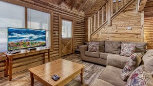 Interno di una baita di legno con pavimento in legno, un divano, un tavolino da caffè e un televisore che mostra un paesaggio montano.