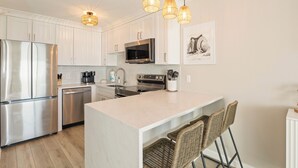 A modern kitchen with white cabinets, stainless steel appliances, a central island with bar seating, and pendant lighting. - Beachfront Beauty - Practically New 1BR Condo! Gulf Front At Perdido Skye (Pensacola)