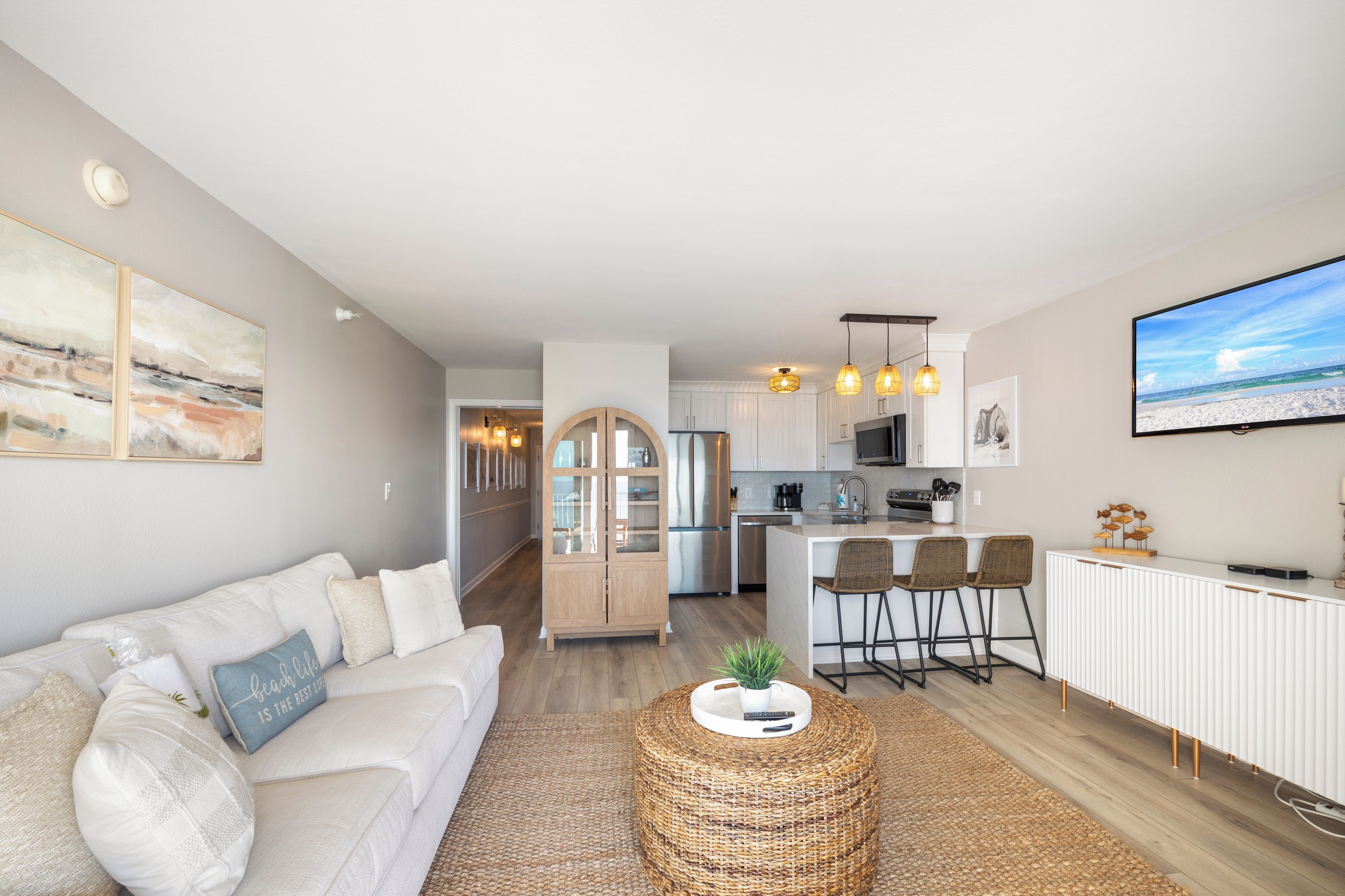 A modern living room with a white sofa, a round wicker coffee table, and a kitchen area with a white countertop and bar stools.