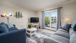 A modern living room with a sofa, a coffee table, a flat-screen TV, and a large window with a view of the outside. - 2-A DELANO CONDO - OCEAN BLOCK-1406 Coastal Hwy, Dewey Beach- DOG FRIENDLY! (Dewey Beach)